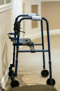 A blue walker with wheels stands beside a wall in a carpeted hallway, suggesting mobility and independence support.