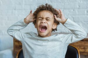 Despaired African American boy with curly hair and closed eyes screaming while standing in light room at home near wall