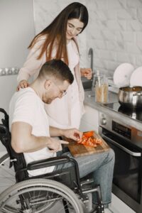 Man in wheelchair chopping vegetables with assistance in a modern kitchen.