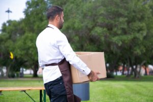 Side view unrecognizable waiter in apron carrying cardboard box in lush summer park